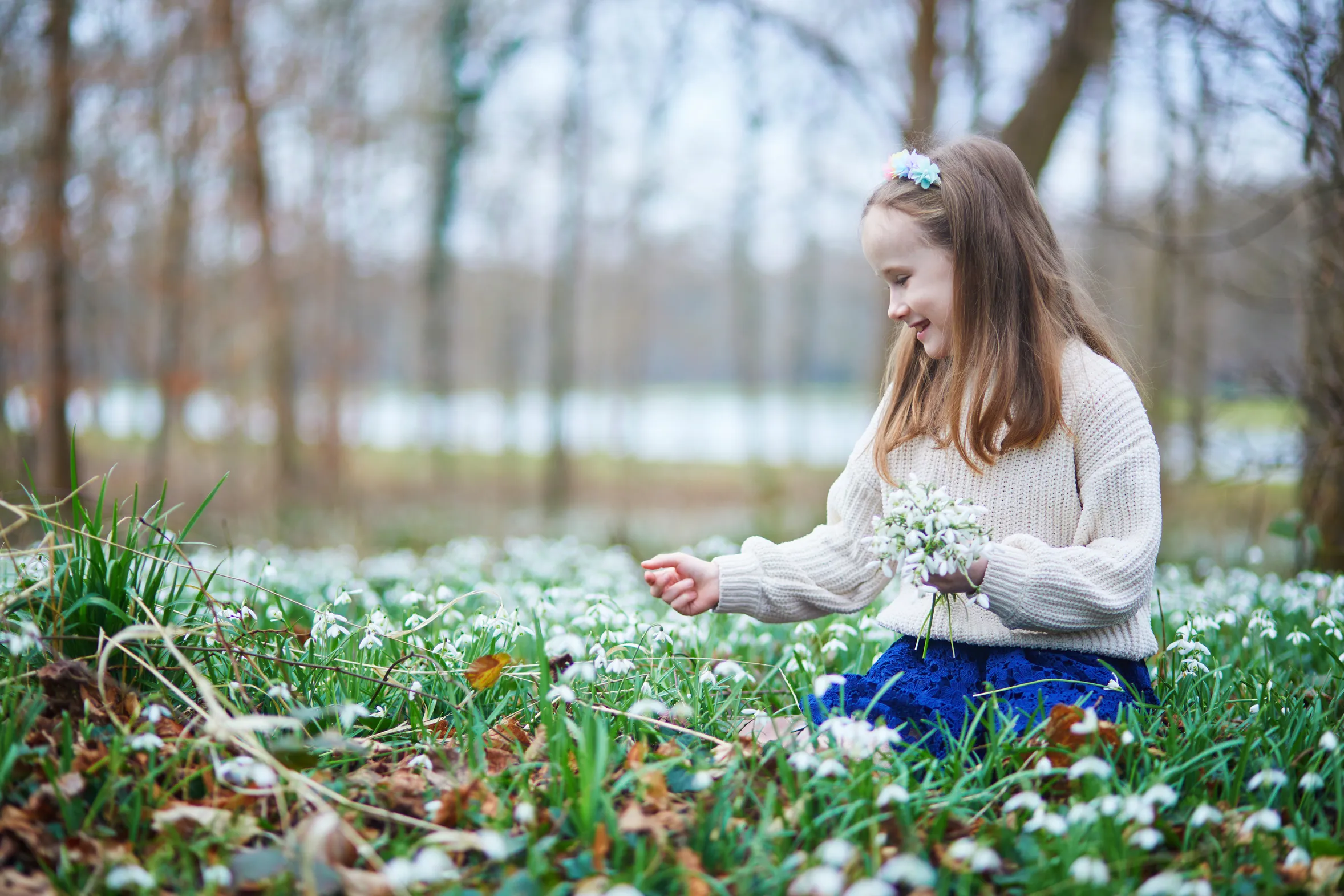 A young girl kneels in a meadow of white snowdrops, smiling as she gathers a small bouquet